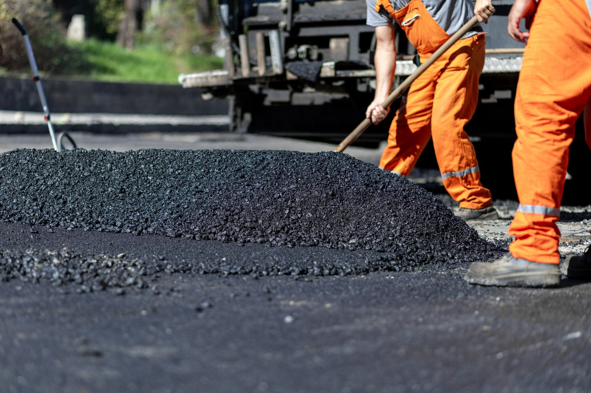 Construction workers in orange overalls are actively spreading hot asphalt on a road